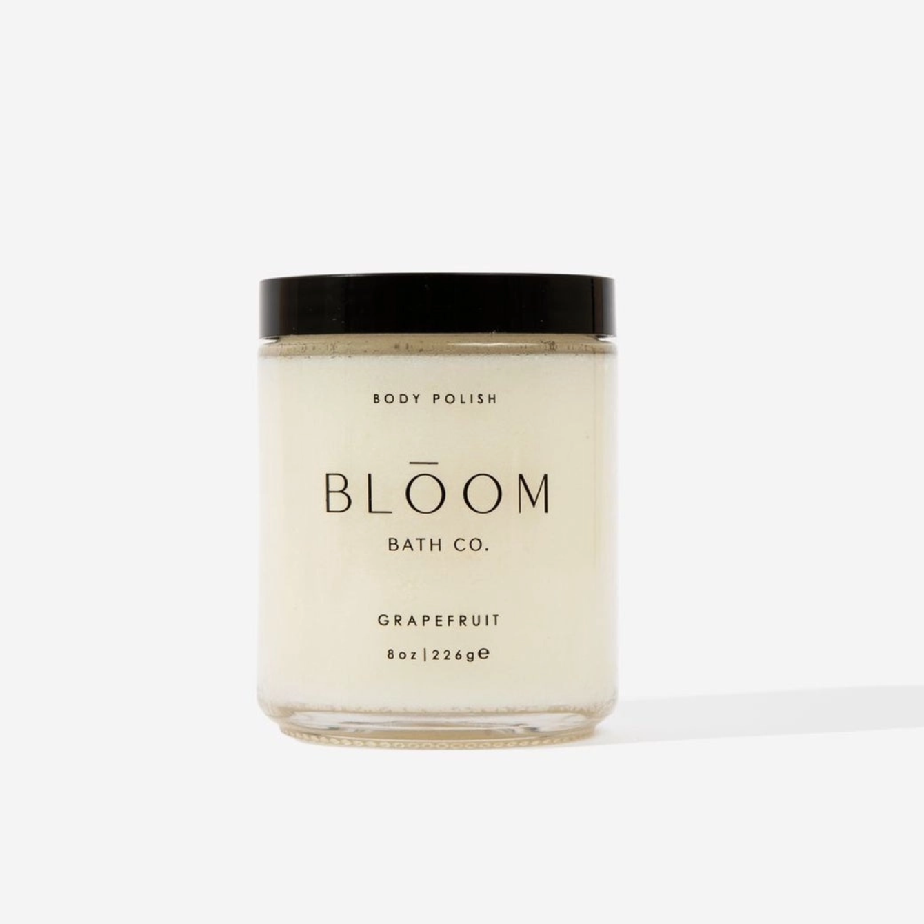 A glass jar of Bloom Bath Co. Grapefruit body polish with a black lid and cream-colored wax, set against a clean white background.