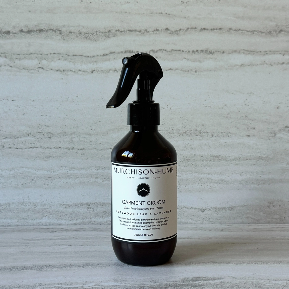 A dark amber glass spray bottle of Murchison-Hume Garment Groom with a black trigger sprayer and a white label, set against a light gray textured background.