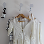 Three white garments hanging on wooden hangers from white wall hooks, with a dark spray bottle on the left hook against a plain white wall.