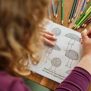 Child coloring a picture of trees with colored pencils on a wooden table.