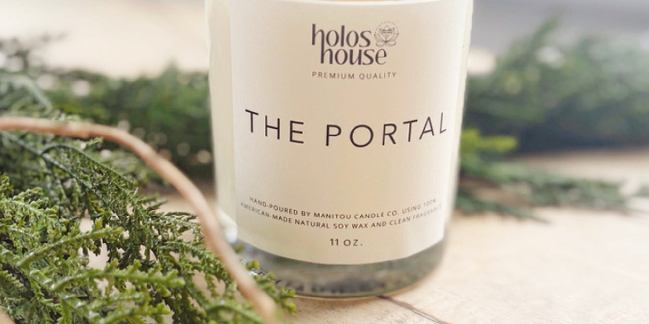Candle with 'holos house' branding and 'The Portal' label on a wooden surface with greenery.