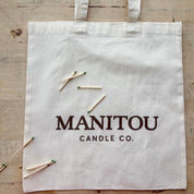 White tote bag with 'Manitou Candle Co.' text and matches on a wooden surface